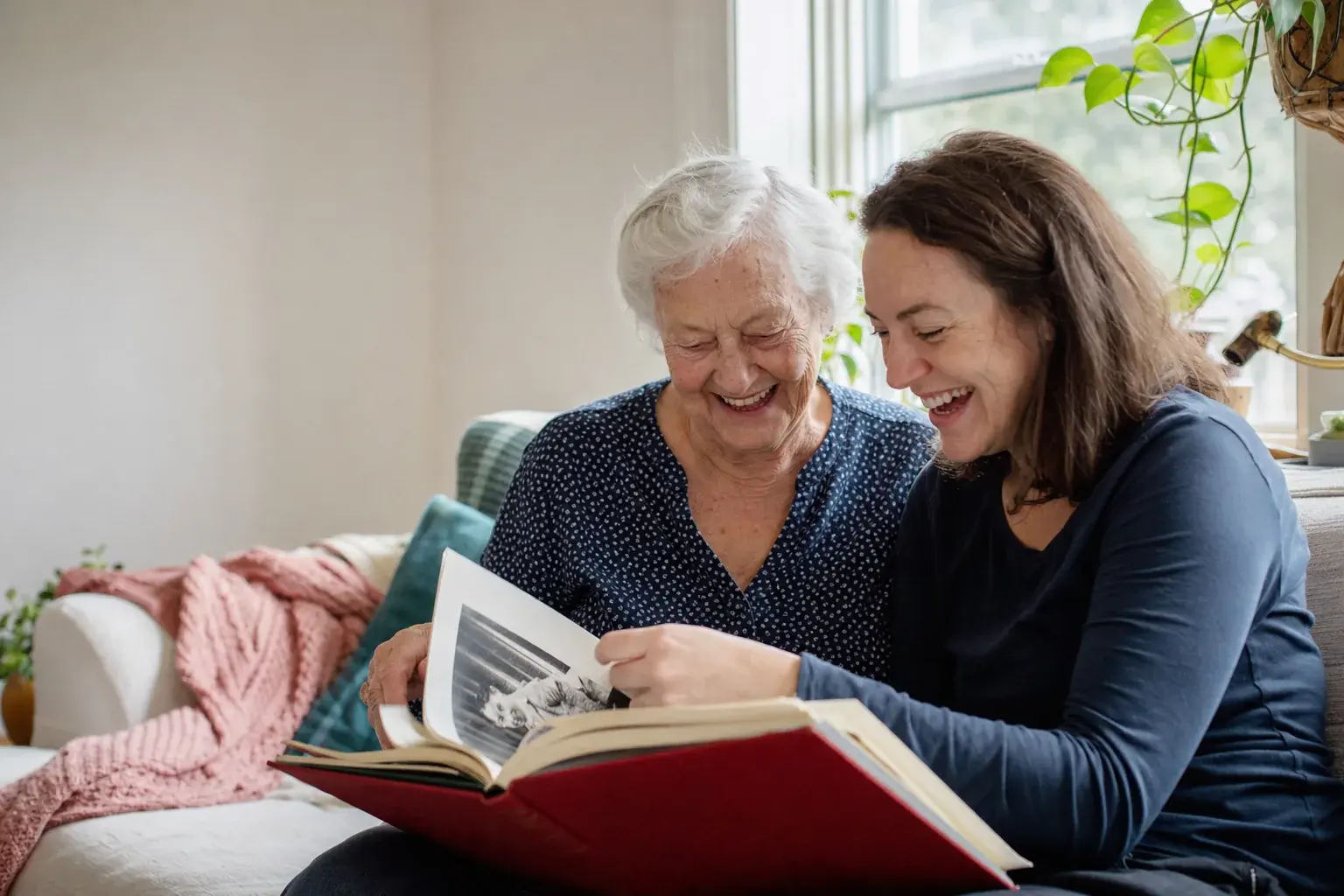 A grandmother and her granddaughter laughing over a hardcover memoir on the sofa